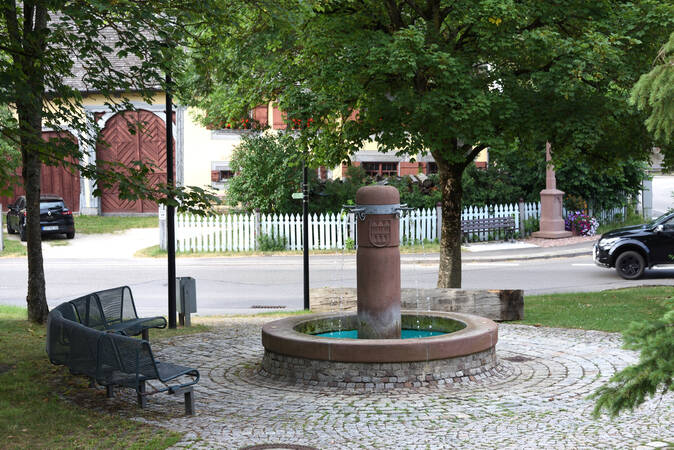 Bild aus der Dorfmitte mit dem mittigen Brunnen, nebenan eine Sitzbank, umrandet von einem Baum